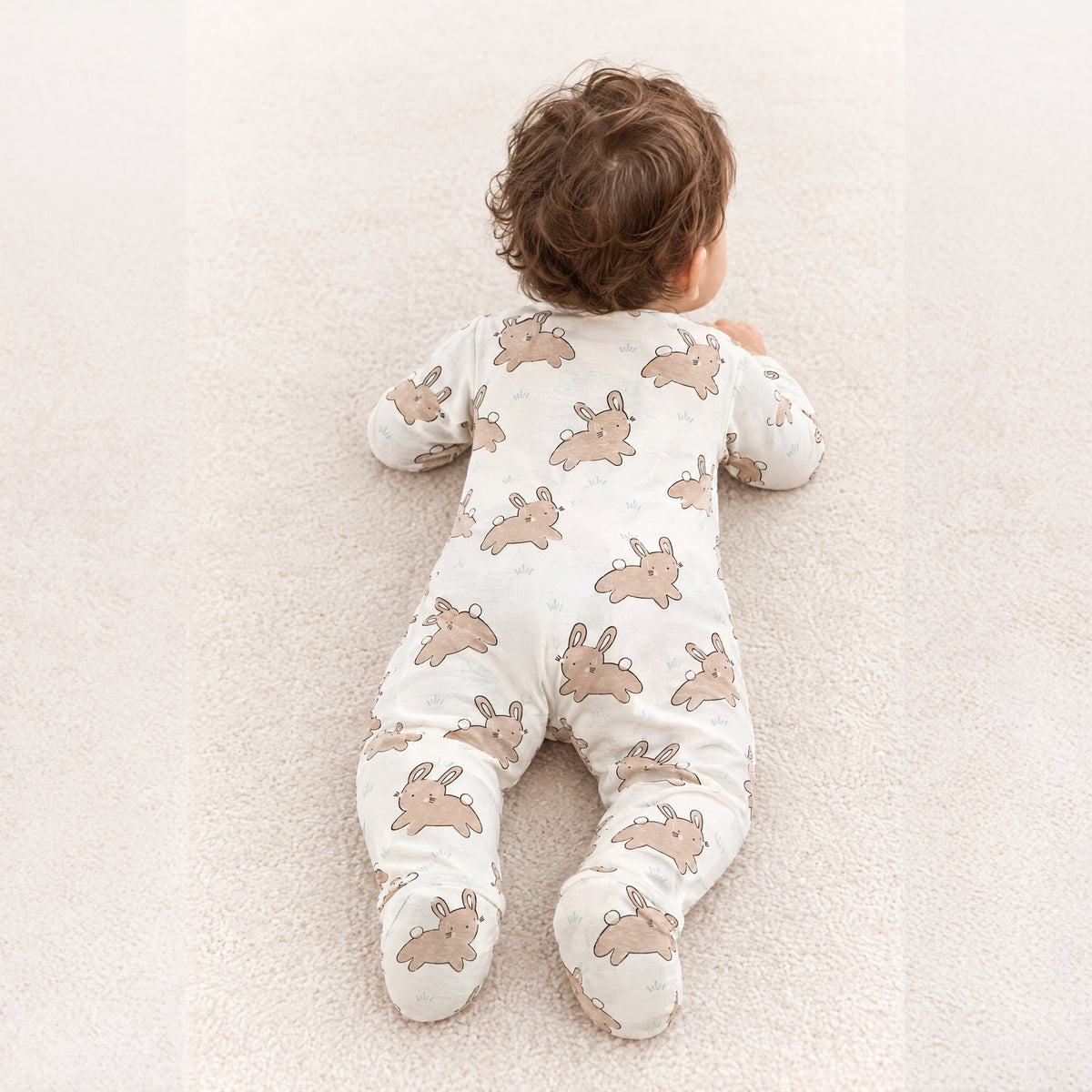 Baby on its tummy on a rug in the cream bunny sleepsuit, back view