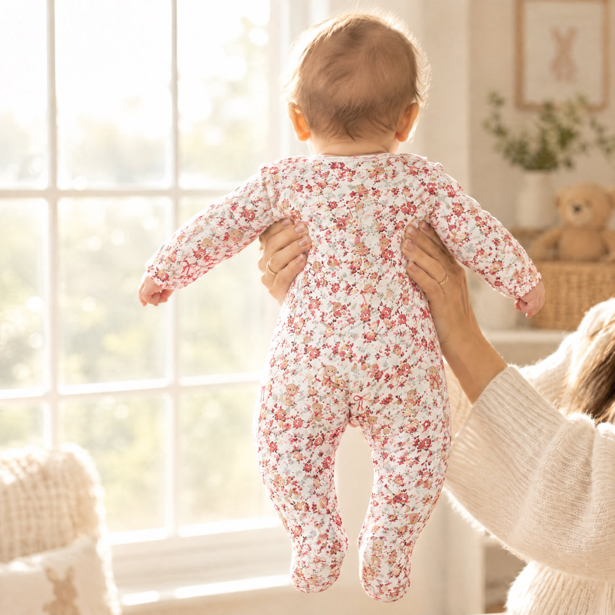 Baby held up by a window in the ivory floral sleepsuit, back view