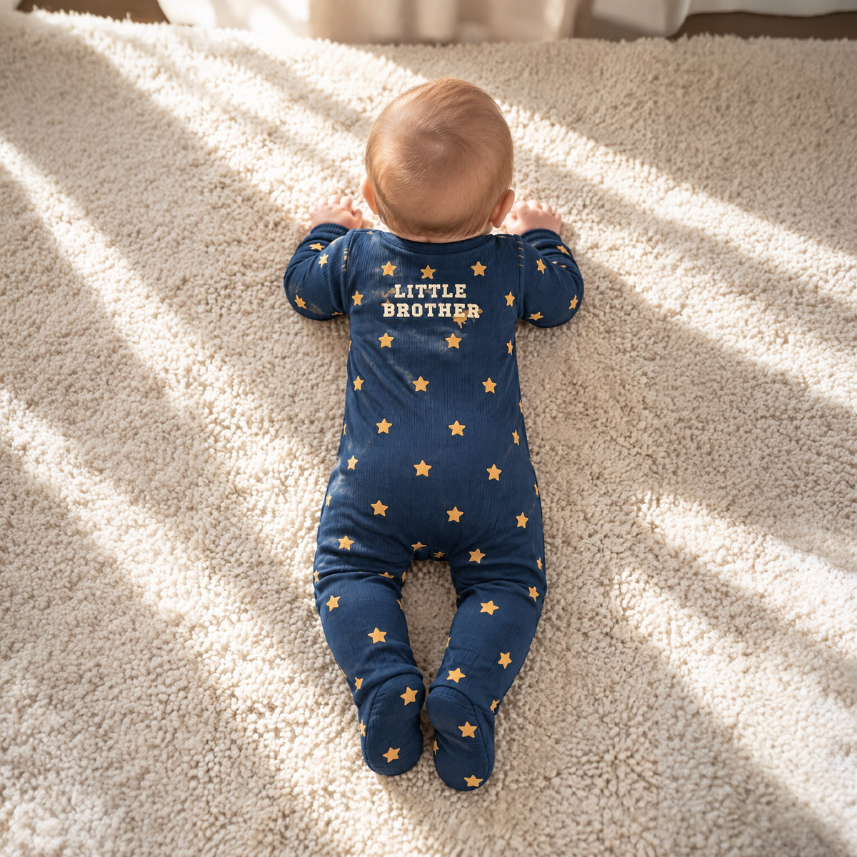 Baby lying on a rug in the navy star sleepsuit, back view with LITTLE BROTHER lettering
