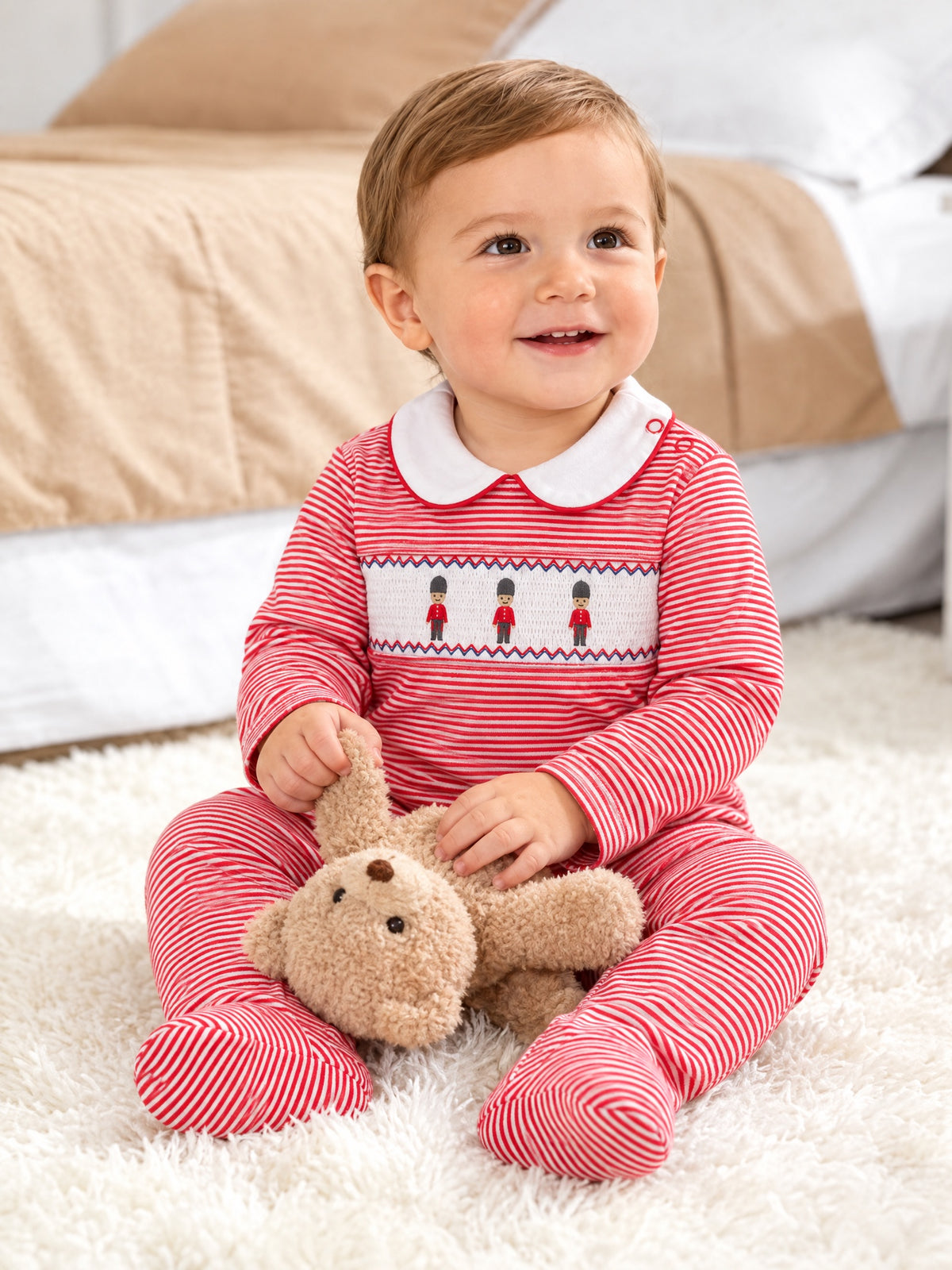 Baby sitting in the red-stripe soldier sleepsuit, front view, holding a teddy